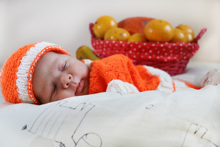 Cute sleeping newborn baby dressed in a knitted orange costume with pumpkin and oranges in a basket behind of him. Autumn halloween or harvest concept.の写真素材