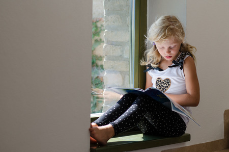 adorable girl with long blond hair sitting in the window table with bookの写真素材