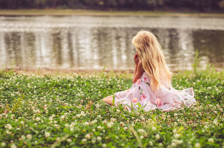 sad lonely girl with long blond hair in pink dress sitting alone as the fairy in the green grass by the riverの写真素材