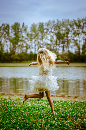 beautiful caucasian girl with long blond hair in colorful dress dancing wild in the green grass by the riverの写真素材