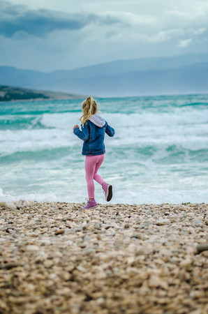 little girl running on the rocky beach with heavy sea with big waves in bad weather の写真素材