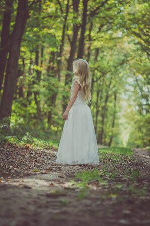 cute little girl in long white wedding dress posing in rural path among autumnal trees in forest in golden hour atmosphereの写真素材