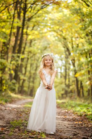 cute little girl in long white wedding dress posing in rural path among autumnal trees in forest in golden hour atmosphereの写真素材