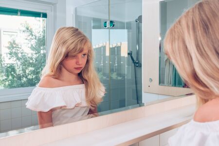 Adorable girl with long blond hair in the bathroom looking into mirror with sad expressionの写真素材