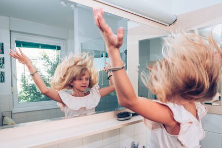Adorable girl in the bathroom looking into mirror and experimenting with her long blond hairの写真素材