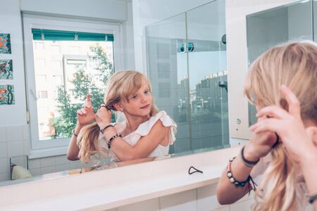 Close up portrait of beautiful girl while doing braid from her long blond hair in the bathroom looking into mirrorの写真素材