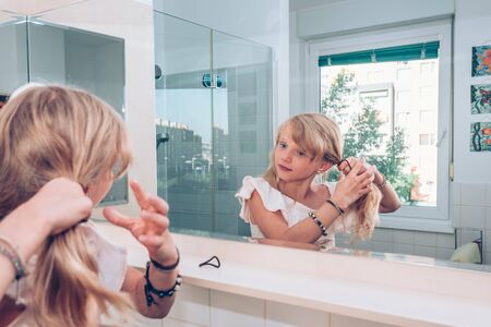 Close up portrait of tidy beautiful girl while doing braid from her long blond hair in the bathroom looking into mirrorの写真素材