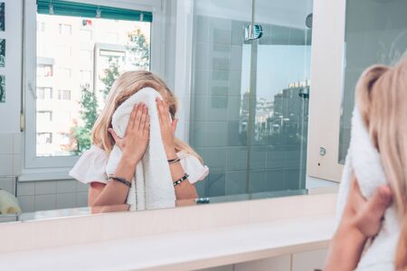 portrait of adorable girl in the bathroom looking into mirror and her face with the towelの写真素材