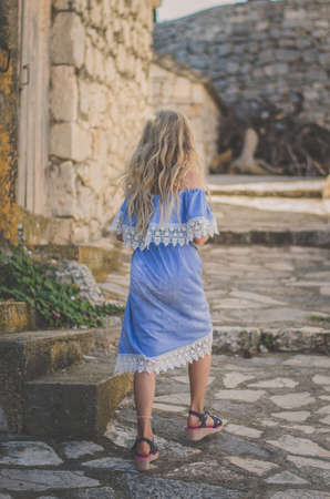 adorable child with long blond hair in blue dress having romantic time in historic streets back viewの写真素材