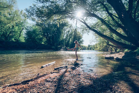 cute girl with long hair back view standing in the river shore relaxing in green scenic nature in sunny golden hour day timeの写真素材