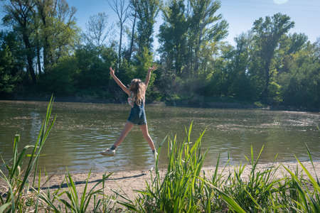 cute girl with long hair back view standing in the river shore relaxing in green scenic nature in sunny golden hour day timeの写真素材