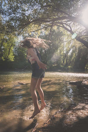 cute girl with long hair back view standing in the river shore relaxing in green scenic nature in sunny golden hour day timeの写真素材