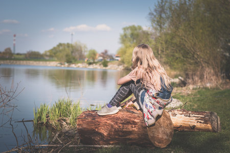 girl with long blond hair sitting by the river in scenic spring nature, back viewの写真素材
