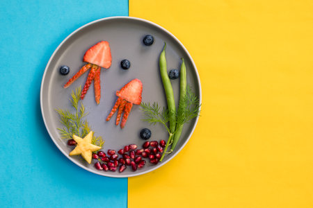 jellyfish, green plants and starfish in sea world from strawberries, blackberries, beans and pomegranate, isolated colorful food ideas on plate with yellow blue backgroundの写真素材