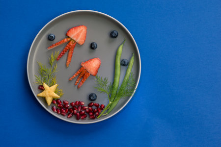 jellyfish, green plants and starfish in sea world from strawberries, blackberries, beans and pomegranate, isolated colorful food ideas on plate with blue backgroundの写真素材