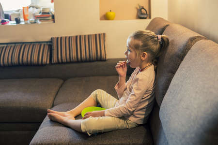 Beautiful little girl eating unhealthy food while watching TV at the sofaの写真素材