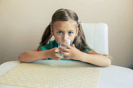 Beautiful little girl drinks clean water from a transparent glass.