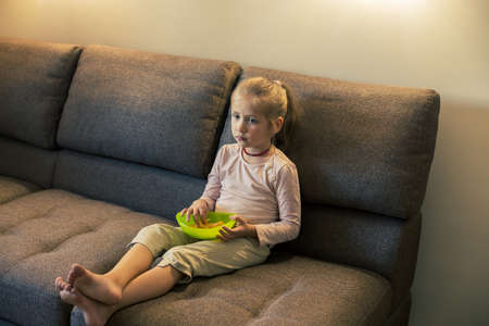 Beautiful little girl eating unhealthy food while watching TV at the sofaの写真素材