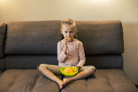 Beautiful little girl eating unhealthy food while watching TV at the sofaの写真素材
