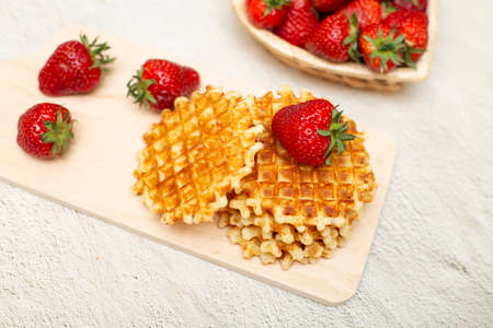 Waffles served with strawberries on a white cutting board on a gray table and a basket with ripe strawberriesの写真素材