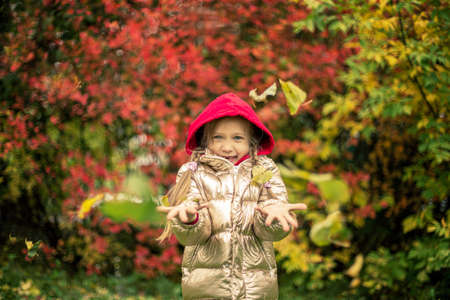 A girl in a gold-colored jacket smiles, throws pink and yellow leaves on an autumn dayの写真素材