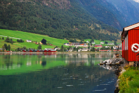 Traditional wooden red cottages on the shores of a fjord in Norway. In the background mountains and houses in a small village. Norwegian landscape.の写真素材