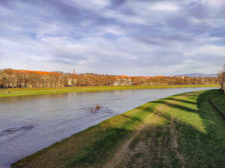 Scenic autumn view of Uzhhorod city and Uzh river, Transcarpathia, Ukraine. Serene riverside landscape captures the gentle flow of water under a cloudy sky alongside a grassy bankの写真素材