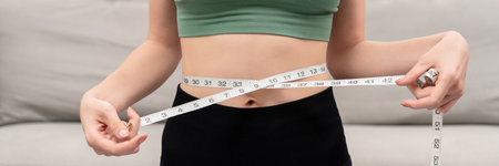 Sporty woman in sportswear is measuring waist circumference and looking on tape measure to checking...