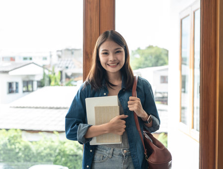 Young woman student is standing and smiling to looking on camera while holding books and bags to preparing for learning lesson classes in college.の写真素材