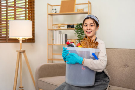 Housekeeper is wearing protective gloves to holding bucket of cleanser bottle and feather duster for working to perform routine house cleaning.の写真素材