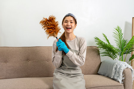 Housekeeper is wearing protective gloves and holding feather duster to cleanups and dusting in living room while working to perform routine house cleaning.の写真素材