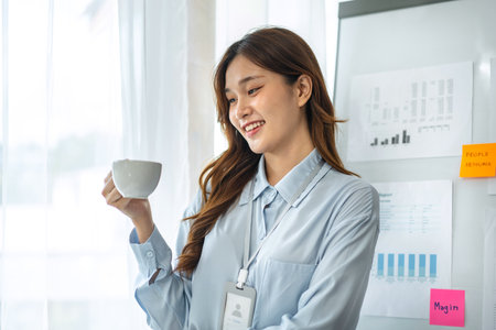Young business woman is standing in front of business information whiteboard and drinking coffee to relaxation after hard work in modern office.の写真素材