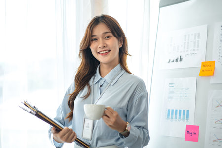 Young business woman is standing in front of business information whiteboard and drinking coffee to relaxation after hard work in modern office.の写真素材