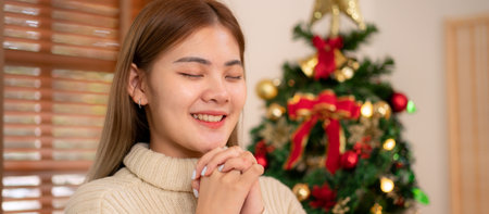 Young asian woman wearing sweater with smiling and praying to god for faith for goodness and forgiveness in cozy living room of christmas decoration at home.の写真素材