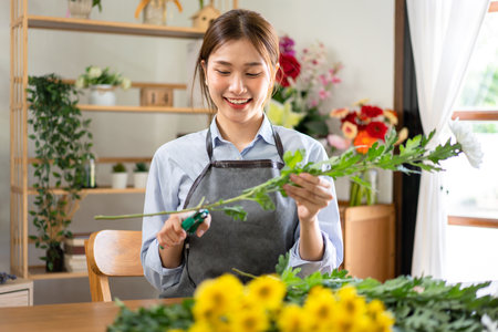 Female florist in apron using scissor to prepare and cutting stem of white chrysanthemum for creating and making flower bouquet in her flower shop.の写真素材