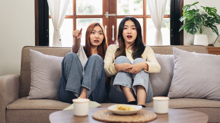 Young woman lesbian couple watching horror movie and pointing with excited face while spending time to relaxation together on big comfortable sofa at home.の写真素材