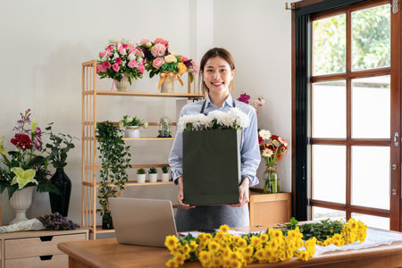Female florist in apron holding paper bag of white chrysanthemum after creating and designing floral for arrangement flower bouquet in her flower shop.の写真素材