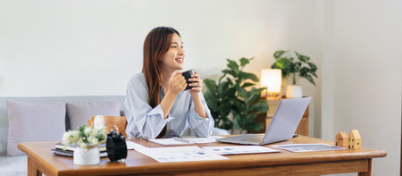 Female freelance is holding coffee to drinking and looking outside to take a break while working on the table in living room at home office.の写真素材