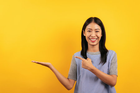 Young asian woman in casual clothes pointing to advertising something on hand with enjoying and smiling with having happiness isolated over yellow background.の写真素材