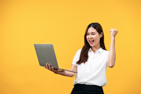 Young asian woman wearing white short sleeve shirt and holding laptop to working while smiling and raising one arm to celebrate after successfully work isolated over yellow background.の写真素材