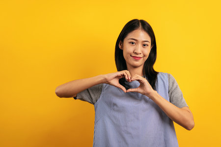 Young asian woman in casual clothes smiling and using hands to doing heart shape feeling love with happiness isolated over yellow background.の写真素材