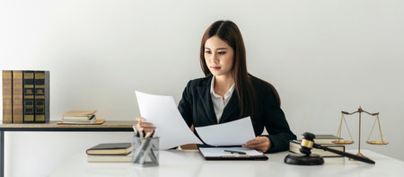 Business lawyer woman holding business contract to reading while sitting to working on the table with brass scales and justice hammer in law firm office.の写真素材