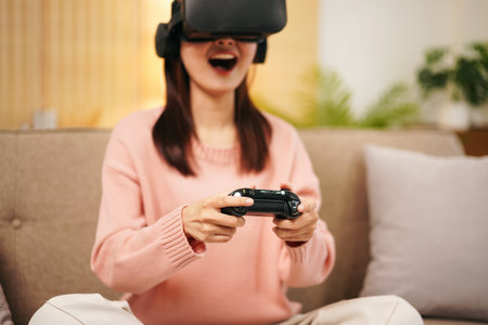 Young woman lesbian wearing sweater and VR goggles while holding joystick to playing video game with experience virtual reality while relaxing time in living room at home.の写真素材
