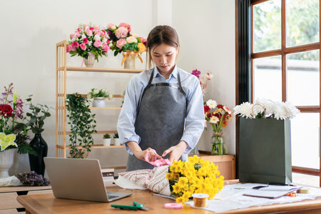 Female florist in apron using craft paper to wrapping yellow chrysanthemum and tie with pink ribbon on the table while creating and designing floral for arrangement flower bouquet in her flower shop.の写真素材