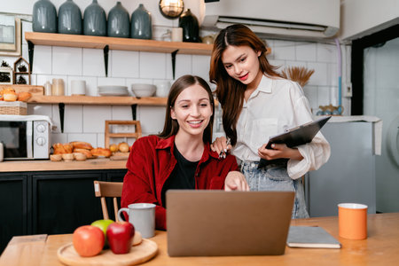 Two young woman pointing data on laptop and reading business information to brainstorming about new project business while working together in the kitchen at home.の写真素材