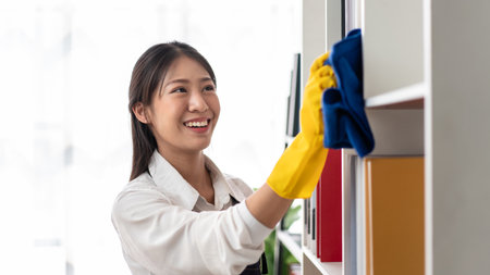 Housewife in apron wearing gloves to using microfiber fabric to wiping cleanup bookshelf and furniture while working and cleaning in the house.の写真素材
