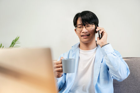 Businessman in casual is sitting comfortable sofa and using smartphone to talking with colleague while drinking hot chocolate and working about business project in laptop at home.の写真素材