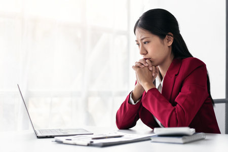 Businesswoman in suit feeling stressed while thoughtful about accounting of new business and typing information with keyboard on laptop during working on the table in modern workspace.の写真素材