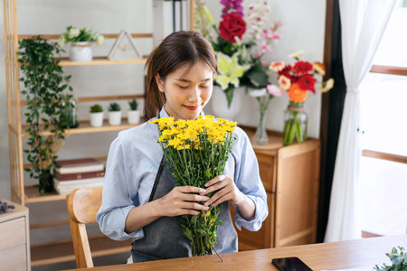 Female florist in apron holding yellow chrysanthemum and smell the flower with freshness while creating and designing floral to arrangement flower bouquet in her flower shop for delivery to customer.の写真素材