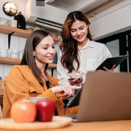Two young woman reading business information in clipboard and digital tablet together to brainstorming about new project business while working with laptop on the table in kitchen at home.の写真素材